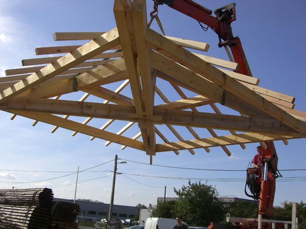 Une grue soulève une charpente en bois sur un chantier, sous un ciel bleu