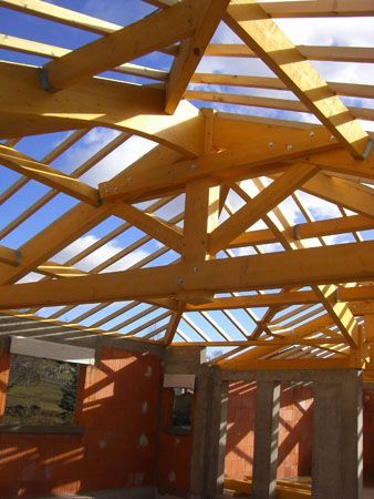 Des fermes de toit en bois apparentes au-dessus d’une structure en briques partiellement construite, sous un ciel bleu