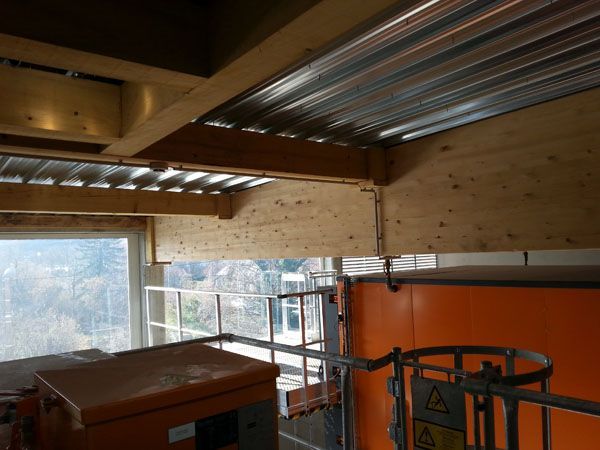 Chantier intérieur avec plafond en tôle ondulée, poutres en bois et engin orange près d’une fenêtre