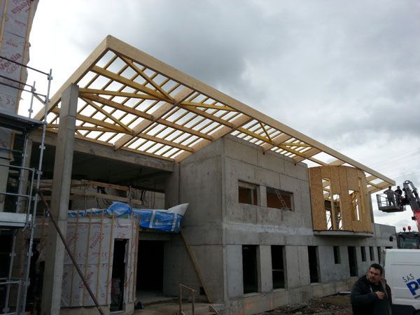 Maison en béton en cours de construction, avec une charpente en bois sous un ciel nuageux et un échafaudage à gauche.