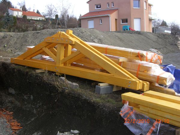 Poutre triangulée en bois jaune sur un chantier, avec des matériaux emballés et des maisons en arrière-plan
