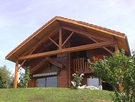 Maison en bois de style chalet, avec un toit pentu, un balcon et des arbustes verts sous un ciel dégagé