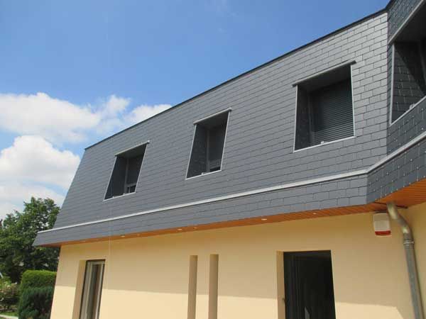 Maison grise à toit en pente avec trois fenêtres en lucarne et un niveau inférieur beige, sous un ciel bleu