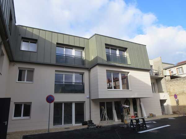 Immeuble moderne de trois étages, avec une façade grise et de grandes fenêtres, sous un ciel partiellement nuageux