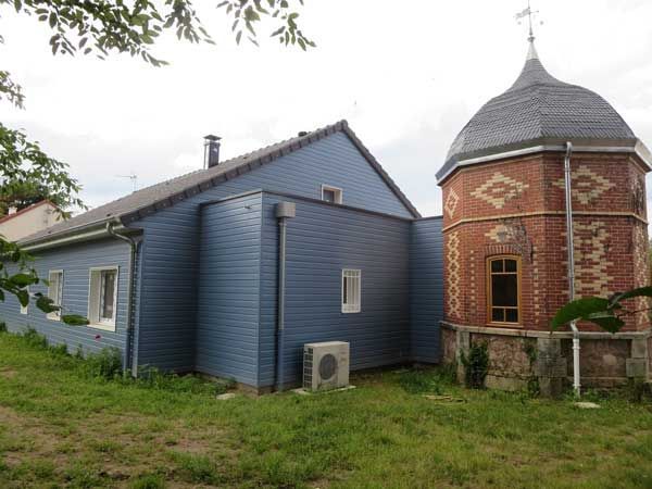 Maison bleue à côté d’une tour ronde en briques rouges, coiffée d’un toit en dôme gris, dans une cour herbeuse