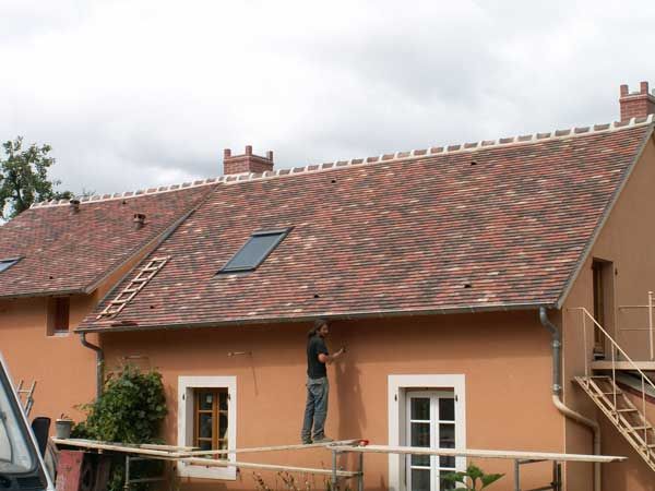 Des ouvriers sur un échafaudage peignent la façade d’une maison couleur pêche au toit de tuiles rouges.