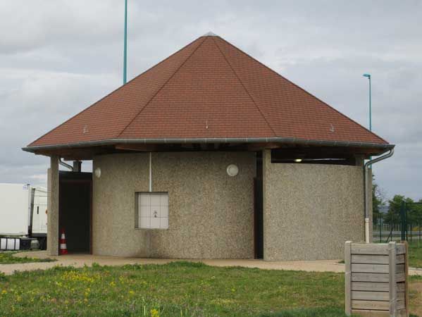 Petit bâtiment utilitaire avec un toit pyramidal rouge, sur un terrain herbeux sous un ciel nuageux.