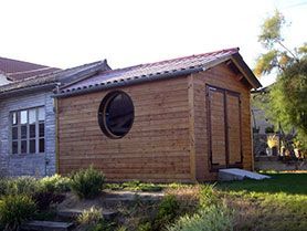 Petit abri de jardin en bois avec une fenêtre ronde et une double porte, à côté d’une maison en pierre dans un jardin.