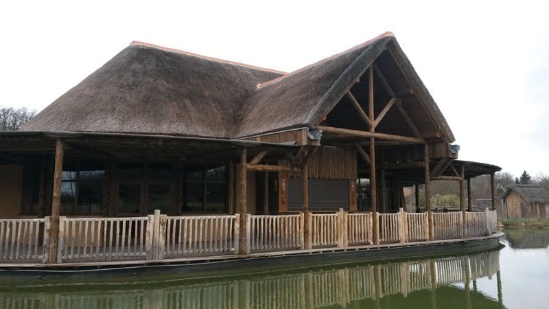 Pavillon en bois au bord d’un lac, avec un toit de chaume et une véranda qui en fait le tour, se reflétant dans une eau calme