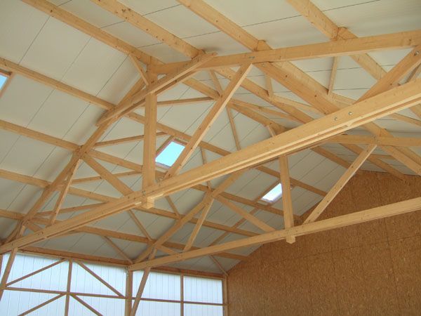 Des fermes et des poutres en bois sous un plafond blanc incliné, à l’intérieur d’une structure de type grange.