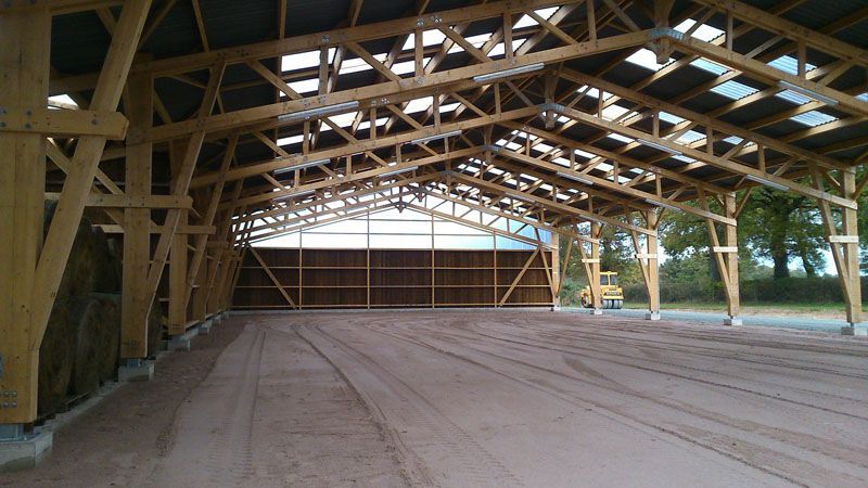 Pavillon couvert en bois, ouvert sur les côtés et doté d’un sol en terre battue, vu de l’intérieur face à un mur fermé.