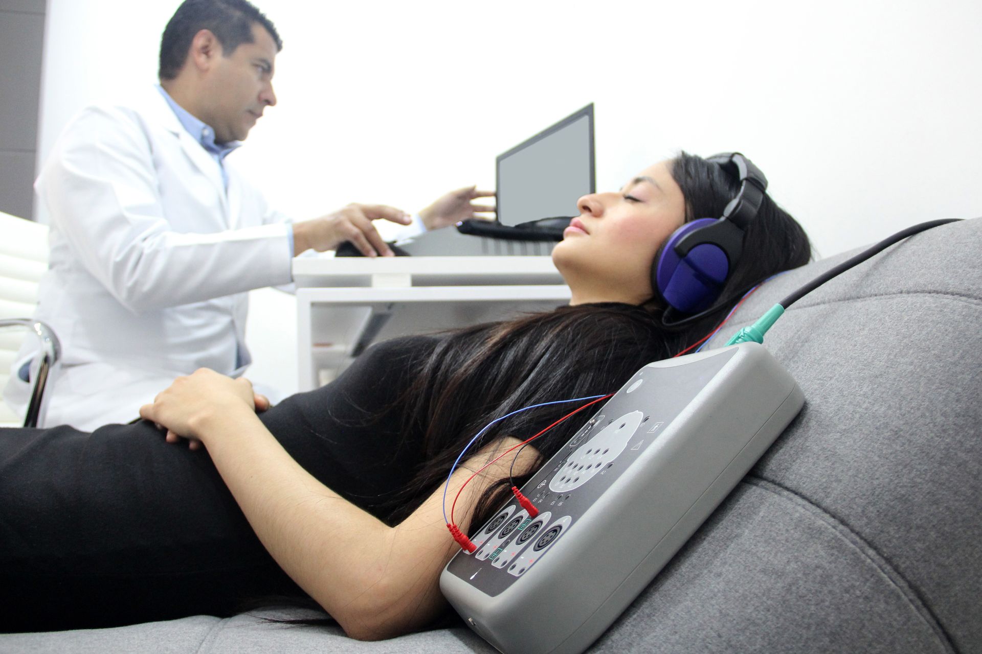 Une femme subit un examen médical avec des écouteurs, reliée à une machine, tandis qu'un médecin utilise un ordinateur portable.