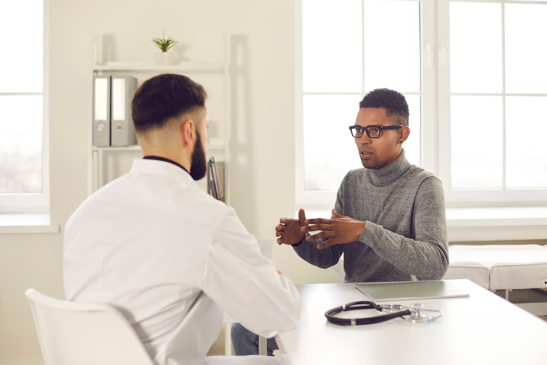 Un patient portant des lunettes consulte un médecin en blouse blanche, la conversation se déroulant à un bureau dans un cabinet médical.
