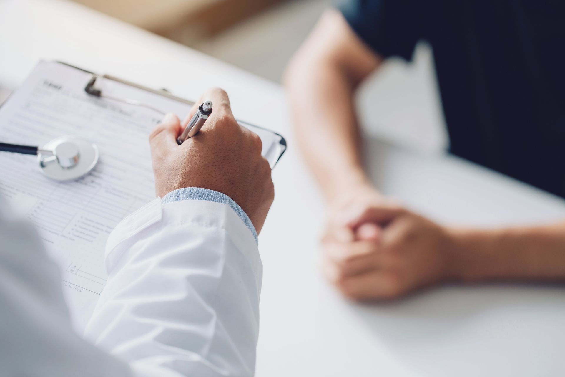 Le médecin écrit sur un bloc-notes, le stéthoscope est posé sur la table ; les mains du patient sont jointes sur une surface blanche.