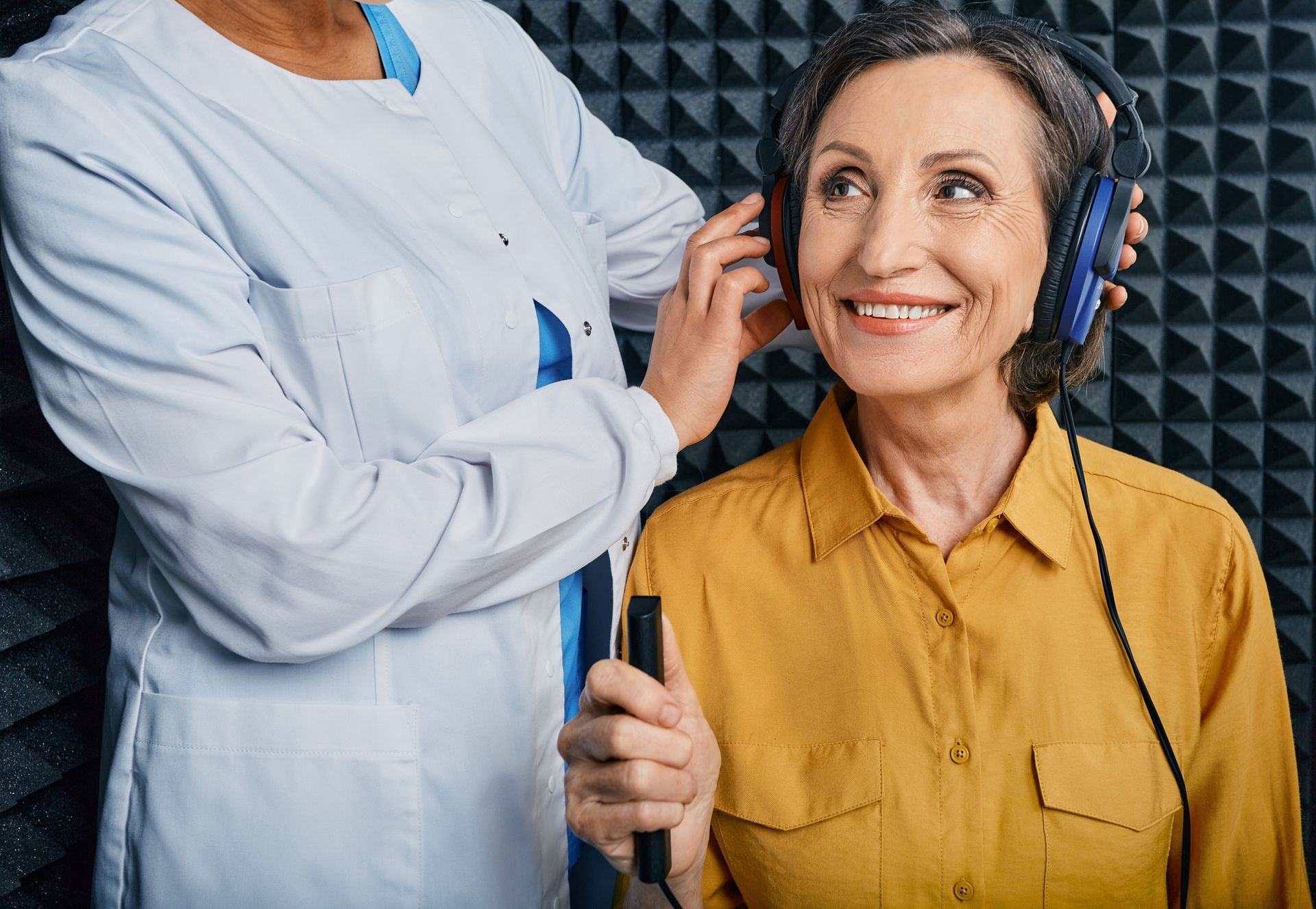 Une femme portant un casque sourit pendant qu'un technicien l'ajuste dans une cabine insonorisée.