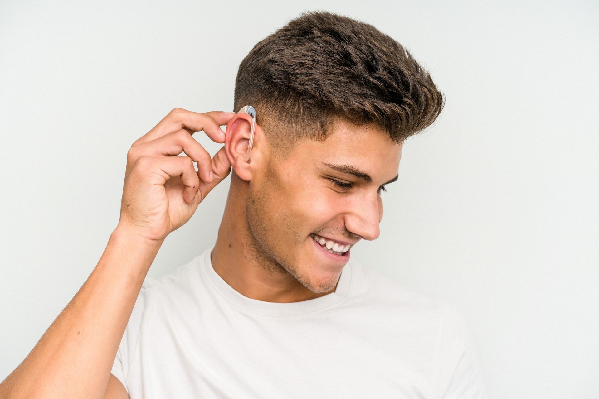 Un homme sourit en ajustant un appareil auditif placé derrière l'oreille. Fond blanc.