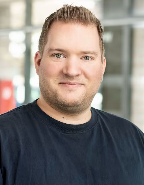 Ein Mann mit Bart trägt ein schwarzes Hemd und lächelt in die Kamera.