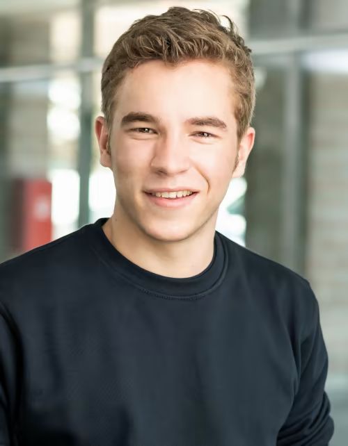 Ein junger Mann in einem schwarzen Pullover lächelt in die Kamera.