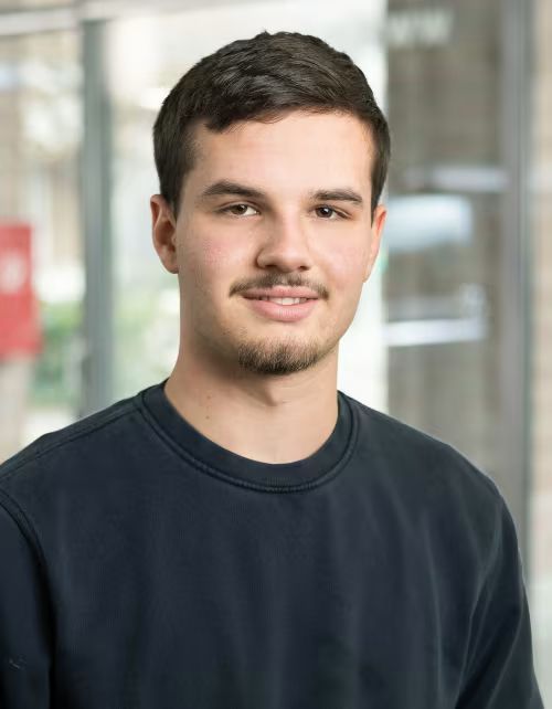Ein junger Mann mit Bart trägt einen schwarzen Pullover und lächelt in die Kamera.