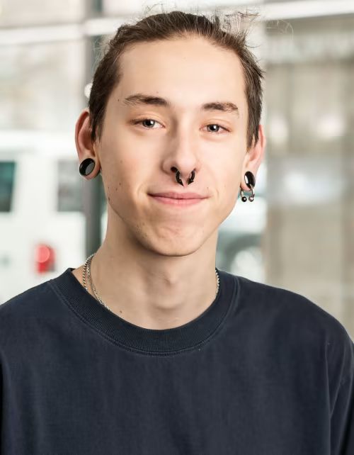 Ein junger Mann mit Nasenring und Ohrstöpseln lächelt in die Kamera.
