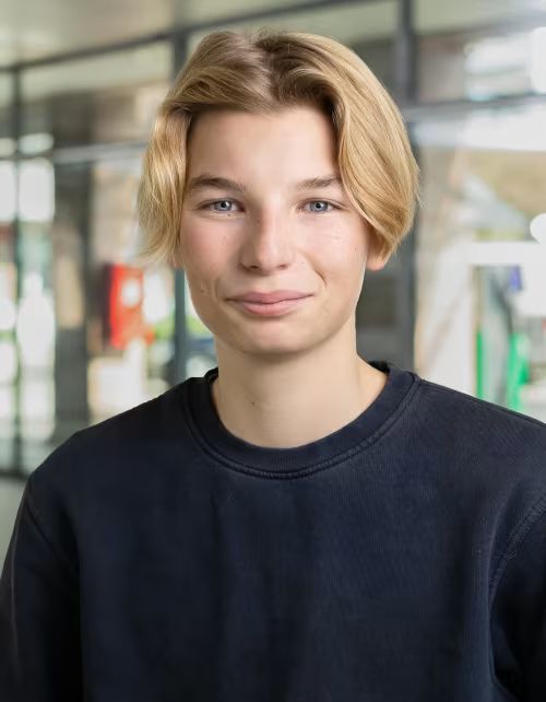 Ein junger Mann mit blondem Haar trägt einen schwarzen Pullover und lächelt in die Kamera.