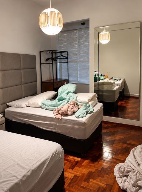 Dormitorio desordenado con la cama sin hacer, un espejo, una lámpara colgante y ropa esparcida por el suelo.