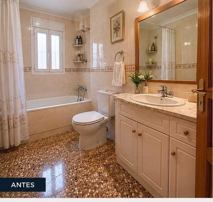 Baño pequeño de color beige con bañera, inodoro, lavabo con mueble, espejo y suelo de baldosas estampadas.