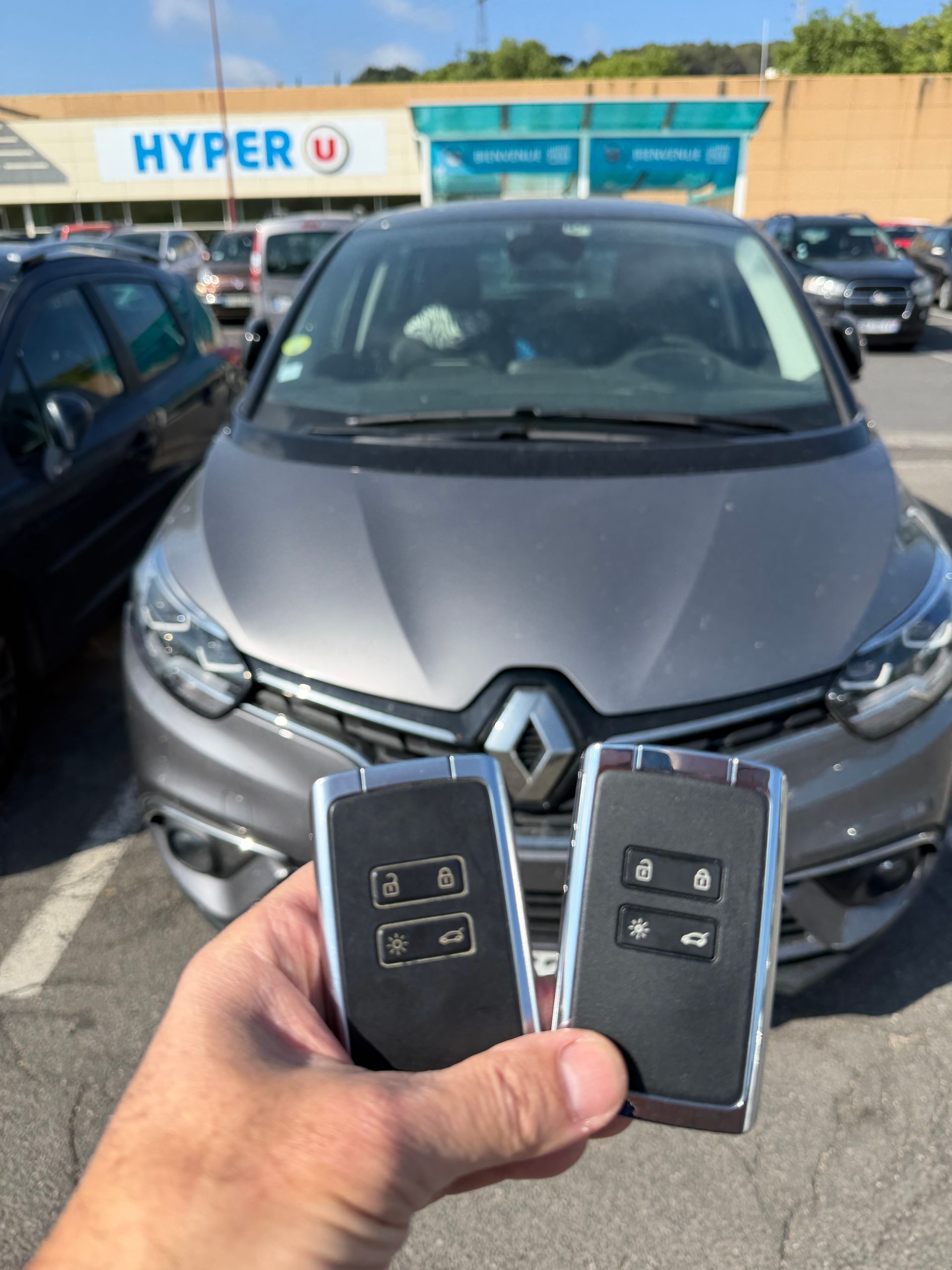 Des mains tenant deux clés Renault devant une Renault grise chez un concessionnaire.
