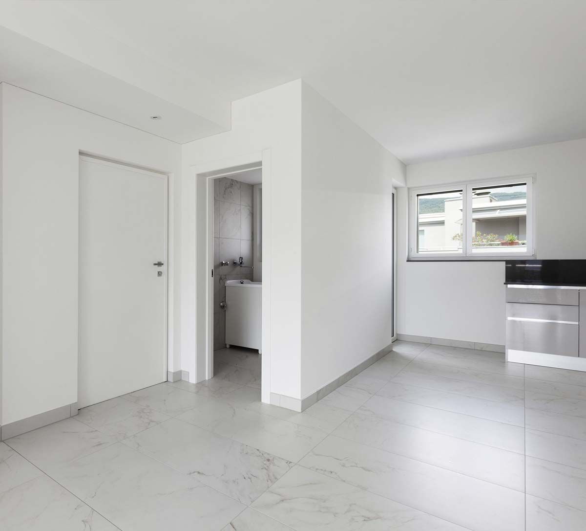 Una habitación vacía con paredes blancas, suelos de baldosas de mármol claro, una puerta blanca cerrada y una pequeña zona de cocina junto a una ventana.