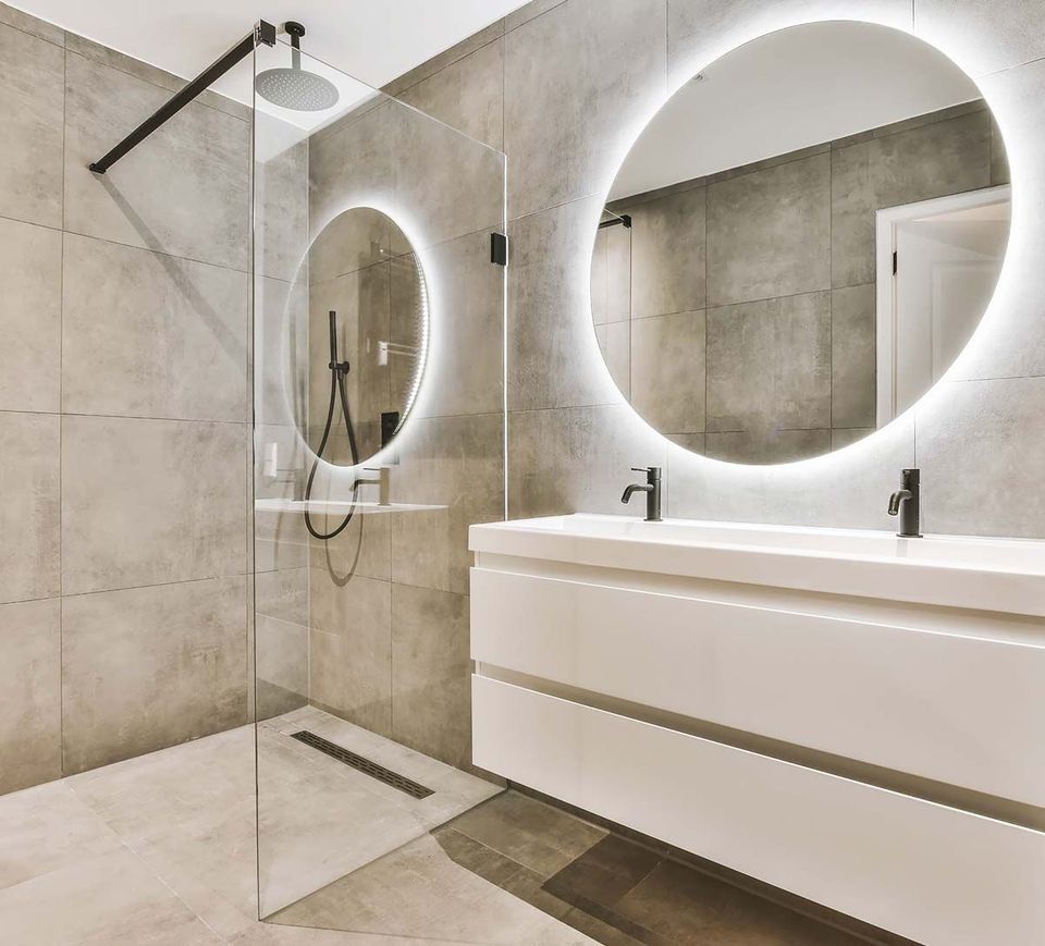 Baño moderno con paredes revestidas de azulejos grises, una ducha acristalada y un tocador doble blanco con espejo redondo retroiluminado.