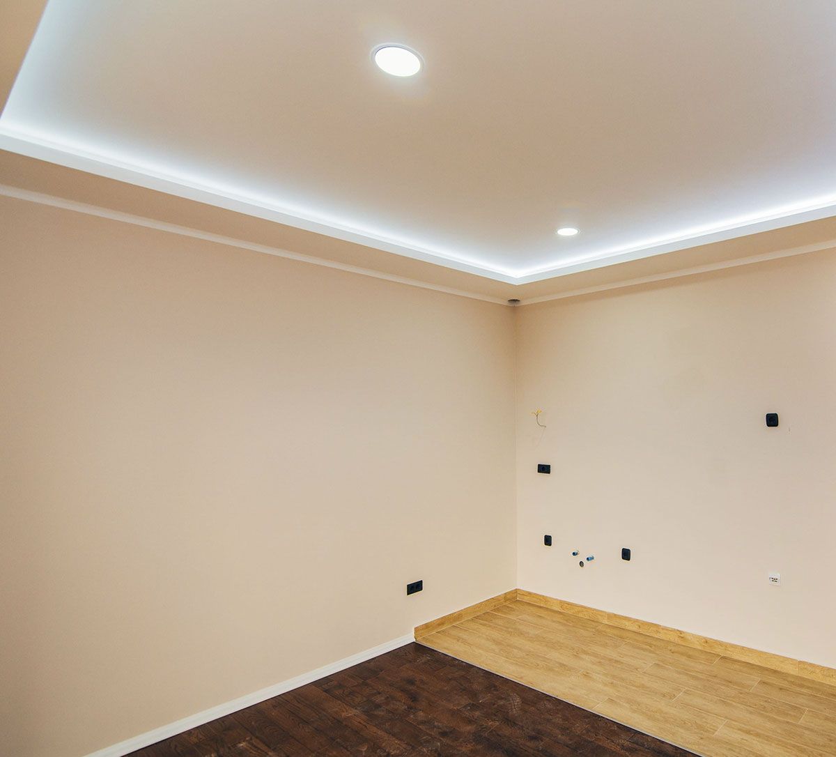 Una habitación vacía con paredes de color beige claro, suelo de madera bicolor (oscuro y claro), techo empotrado e iluminación indirecta.