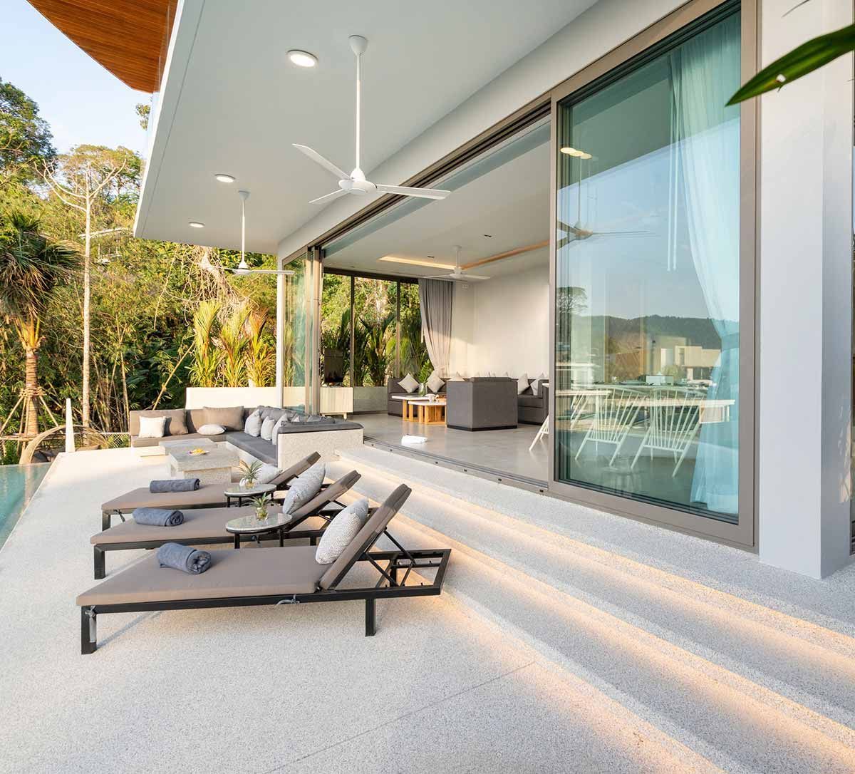 Exterior de una lujosa villa con puertas corredizas de cristal. Tumbonas en una terraza con vistas a la piscina y a una exuberante vegetación.