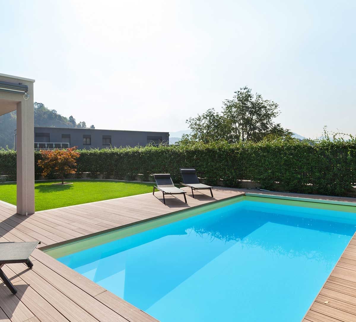 Piscina de agua azul, rodeada de terraza de madera y césped, con tumbonas.