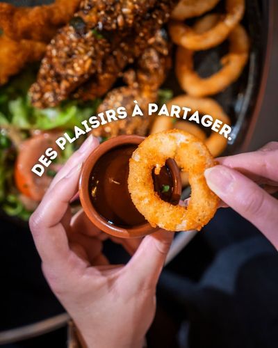 Des mains trempent un rond d’oignon doré dans un petit pot de sauce, à côté d’une assiette de fritures.