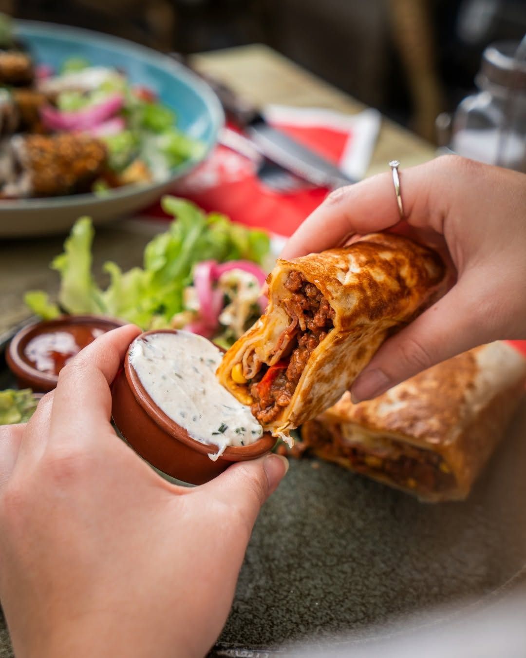 Des mains qui tiennent un burritos et la trempent dans une sauce blanche, à table