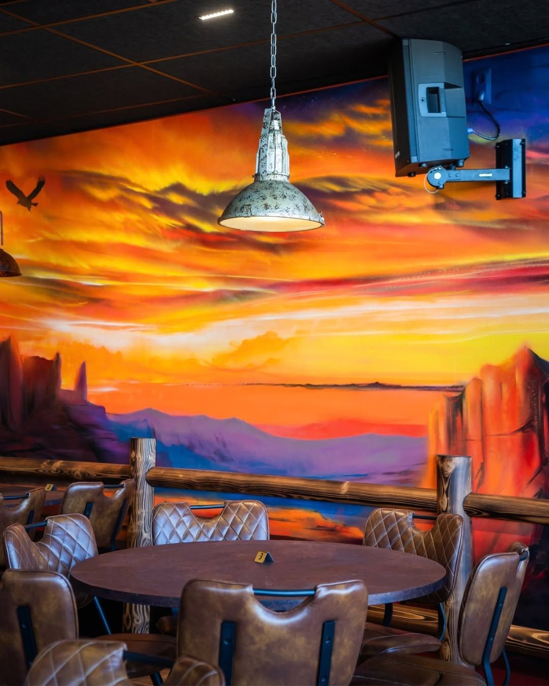 Table ronde dans une pièce rustique, avec une fresque représentant un coucher de soleil éclatant sur le mur et une suspension au-dessus.