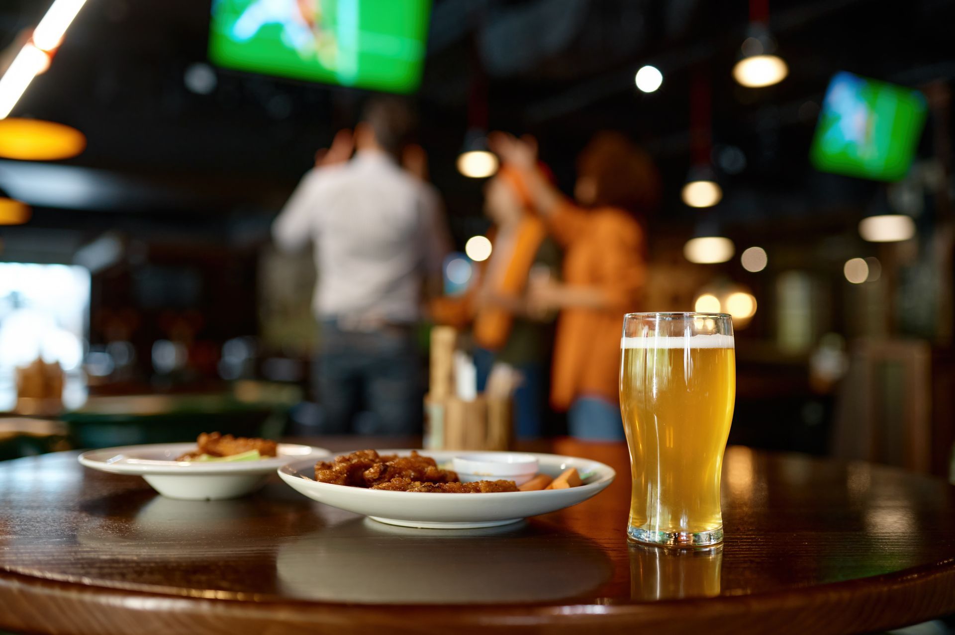 Un verre de bière sur un comptoir, avec des assiettes garnies et des personnes floues regardant la télévision en arrière-plan