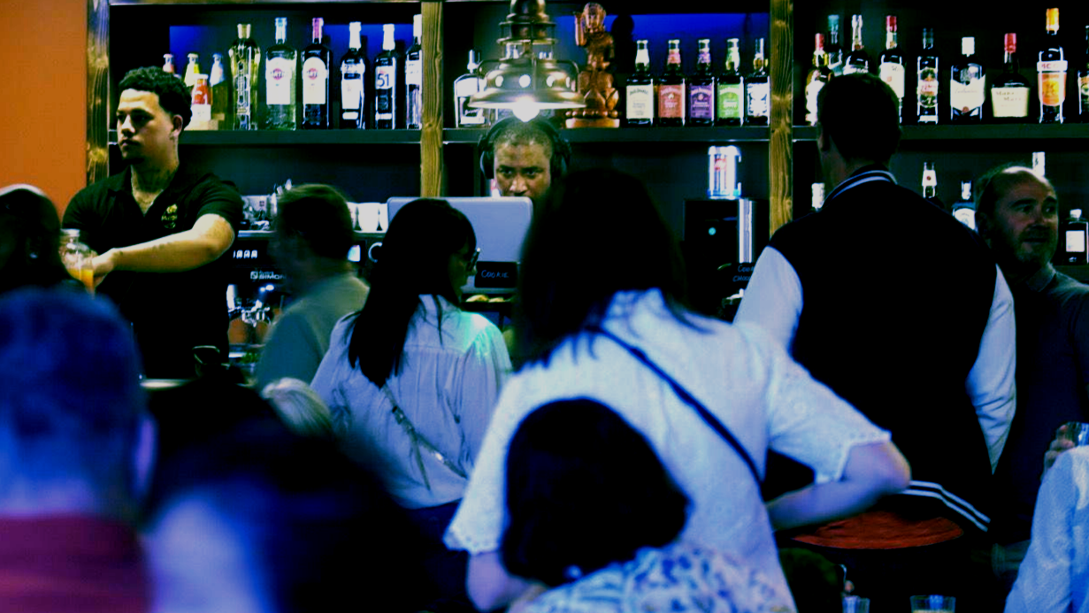 Intérieur sombre d’un bar où des clients sont assis à des tables et un barman se tient derrière des rangées de bouteilles.