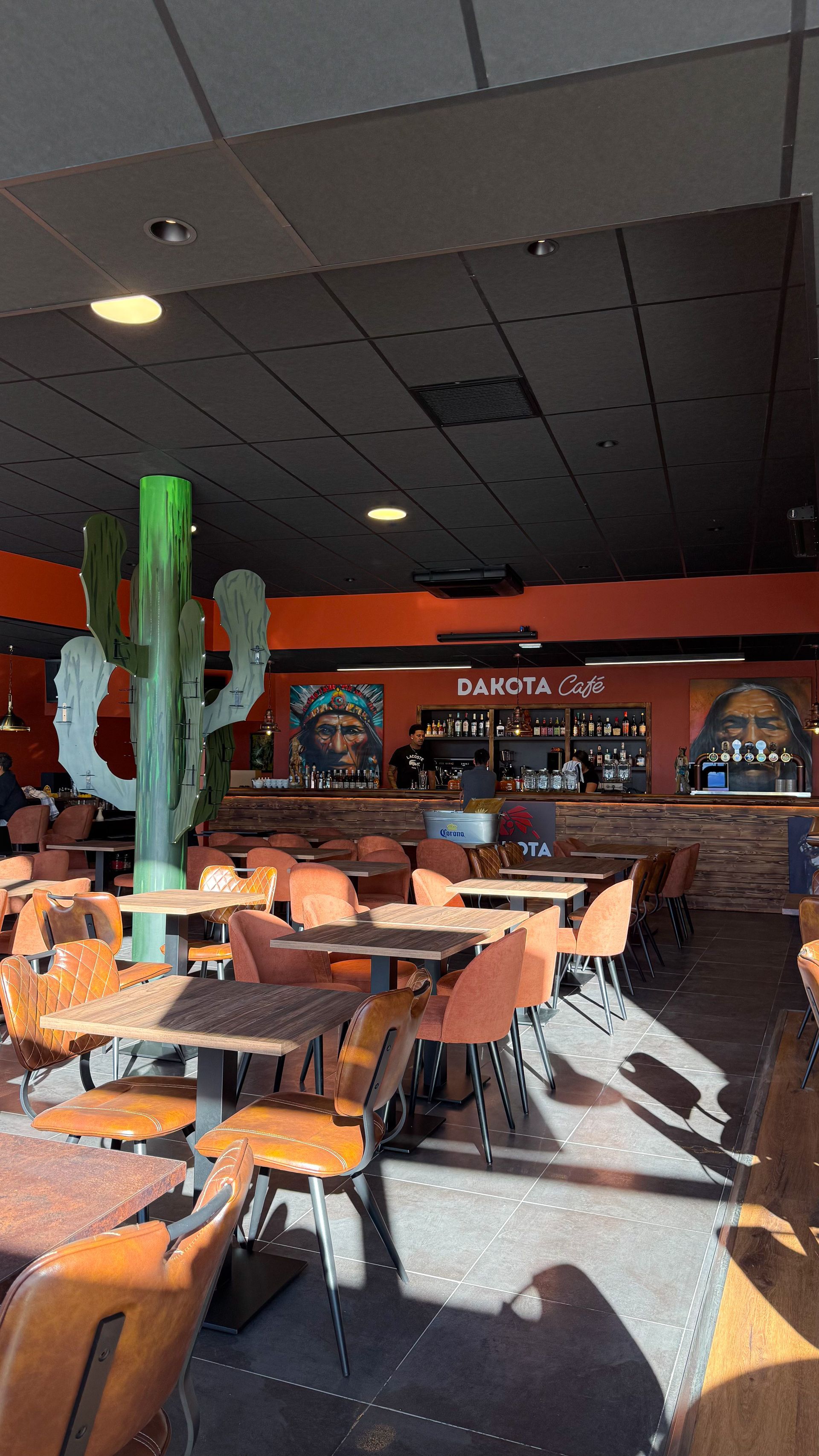 Intérieur de restaurant baigné de soleil, avec des chaises orange, une décoration avec des cactus et un bar en arrière-plan