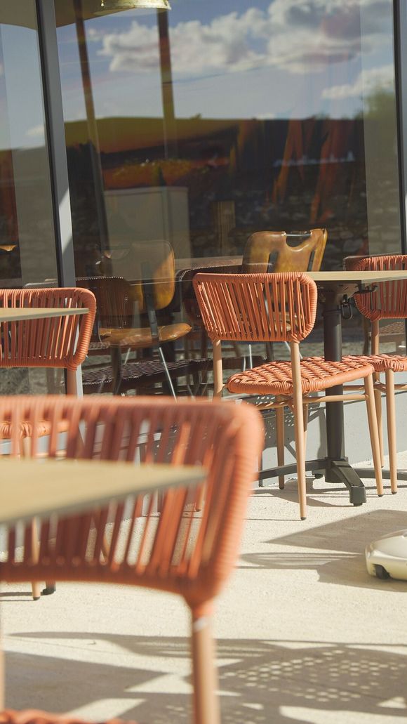 Des chaises et des tables en osier orange sur la terrasse ensoleillée d’un café, avec des fenêtres vitrées en arrière-plan