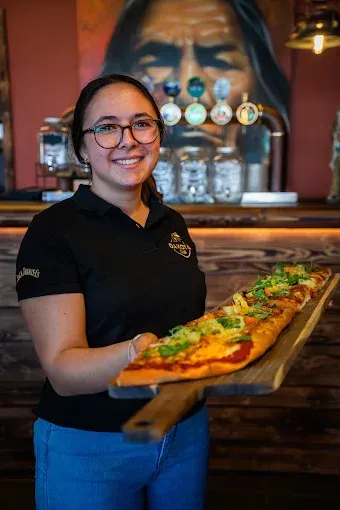 Serveuse souriante en chemise noire tenant un plateau de pizzas dans un bar, avec des tireuses à bière derrière elle
