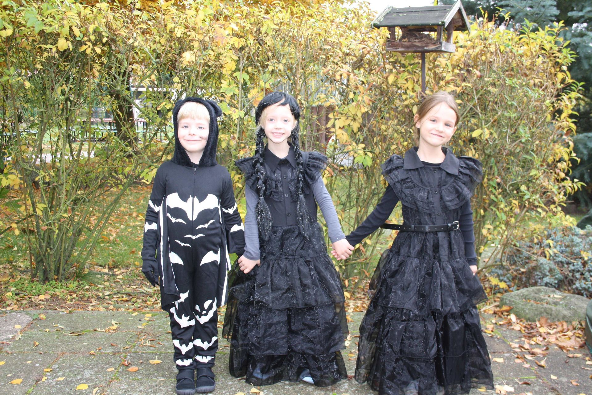 Three children in black costumes are holding hands in front of a birdhouse
