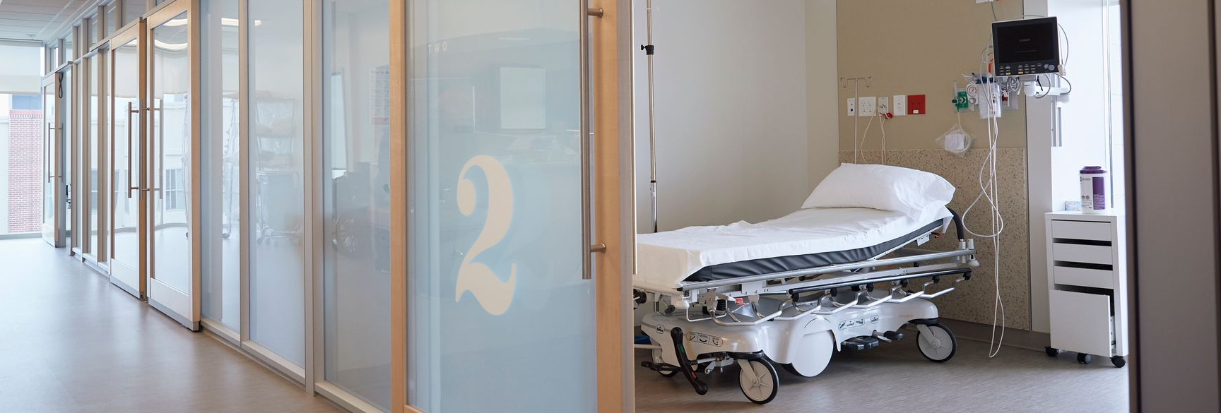 Couloir d'hôpital avec portes en verre dépoli et salle d'examen avec lit médicalisé.