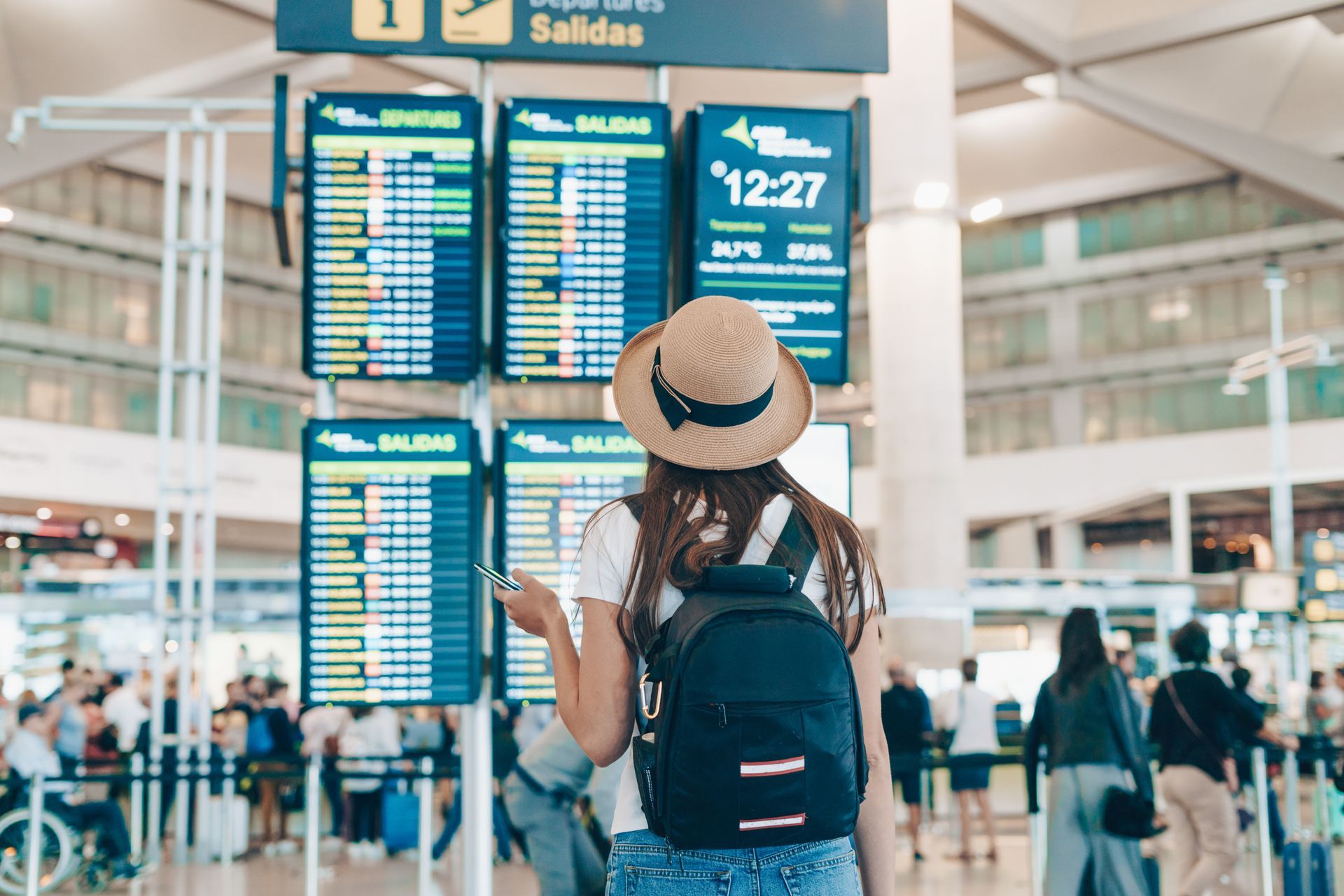 Une femme avec un sac à dos et un chapeau regarde les panneaux d'affichage des départs dans un aéroport.