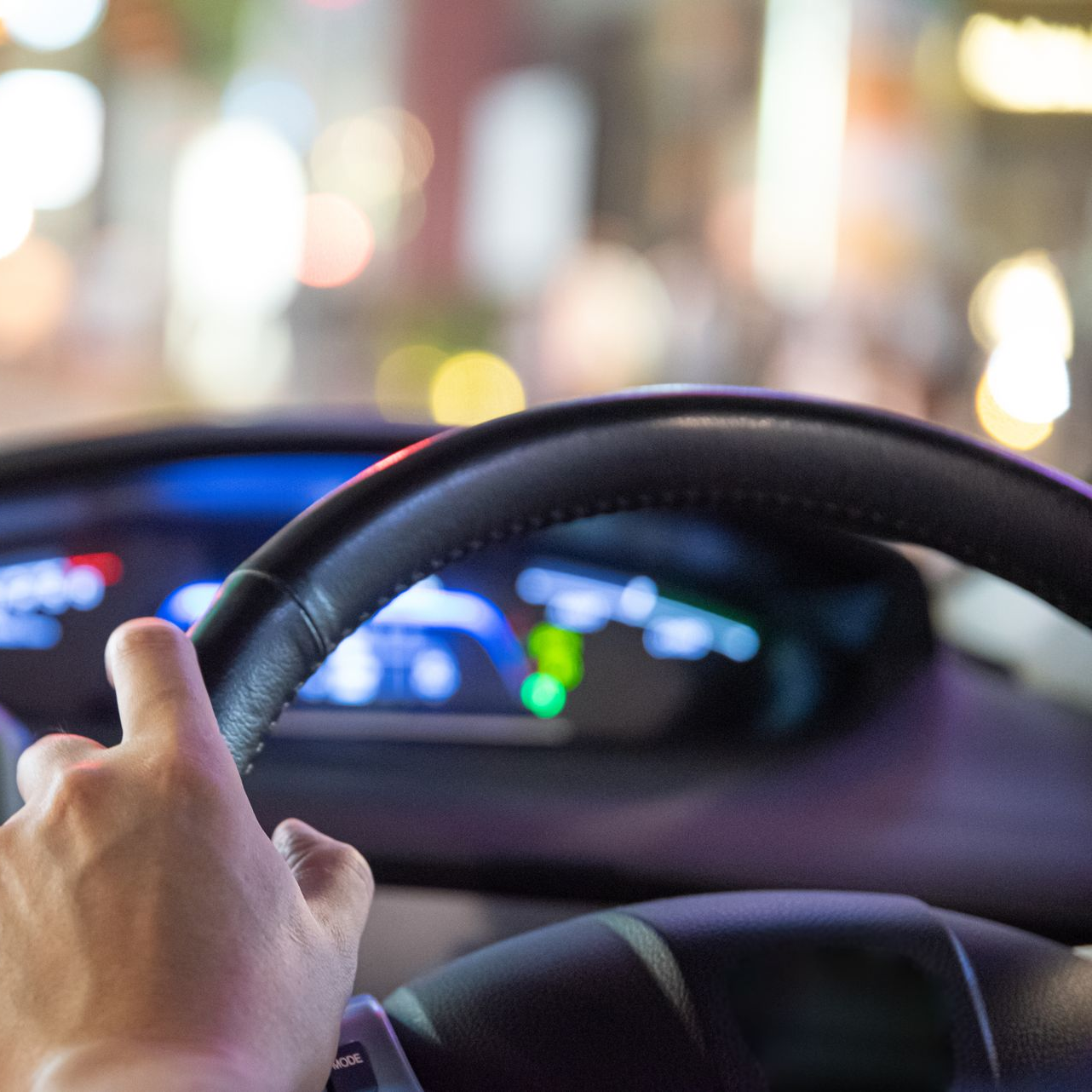 Une main agrippée à un volant noir, conduite de nuit. Tableau de bord et lumières de la ville floues en arrière-plan.