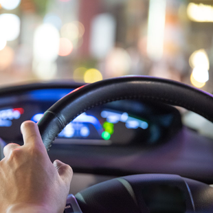 Une main agrippée à un volant noir, conduite de nuit. Tableau de bord et lumières de la ville floues en arrière-plan.