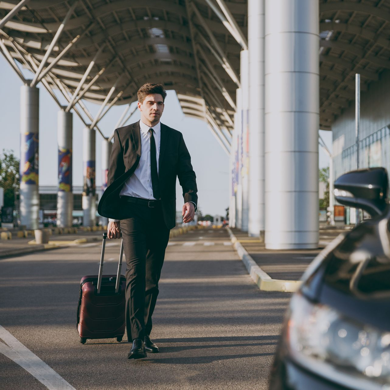 Un homme en costume, une valise à la main, marche vers une voiture noire garée devant un immeuble.