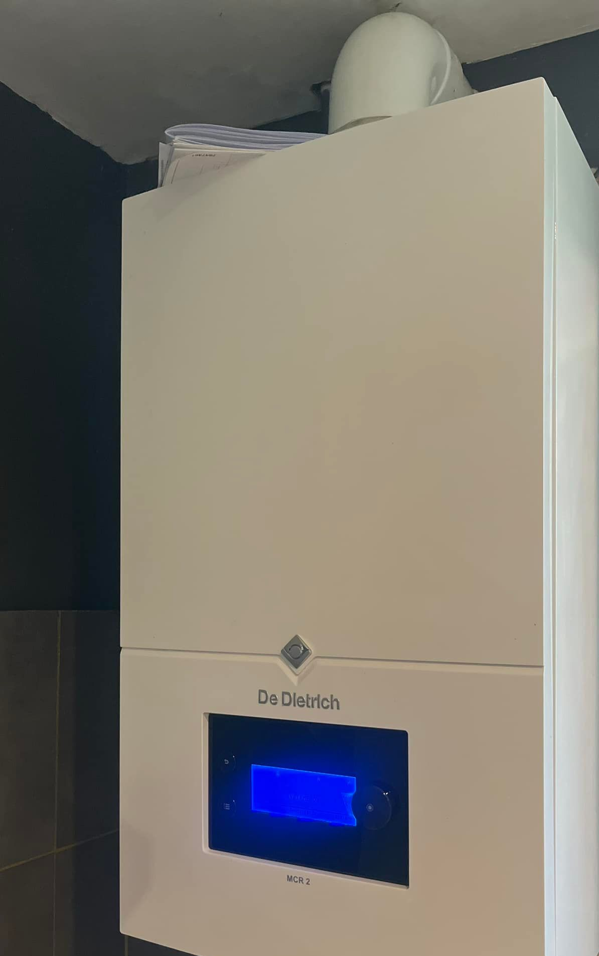 Chaudière à gaz blanche fixée au mur avec affichage numérique bleu ; conduit d'évacuation blanc au-dessus.