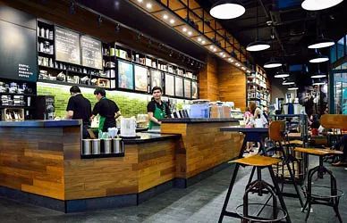 Interior de cafetería moderna con mostrador de madera, baristas, lámparas colgantes y taburetes en las mesas.