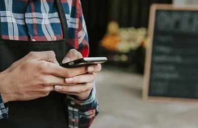 Manos sosteniendo un teléfono inteligente, con un letrero de pizarra borroso al fondo.